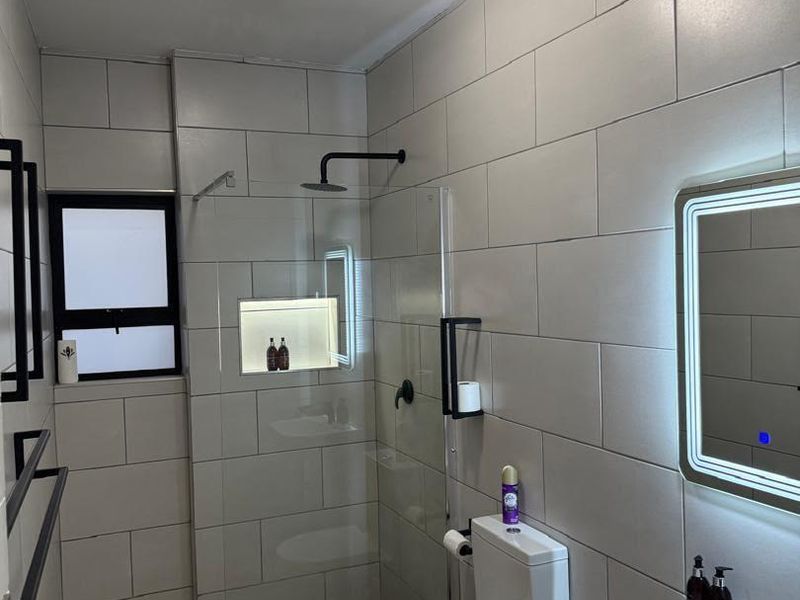 Modern bathroom featuring a walk-in shower with a recessed niche, black fixtures, an illuminated vanity mirror, and contemporary neutral tiling throughout.