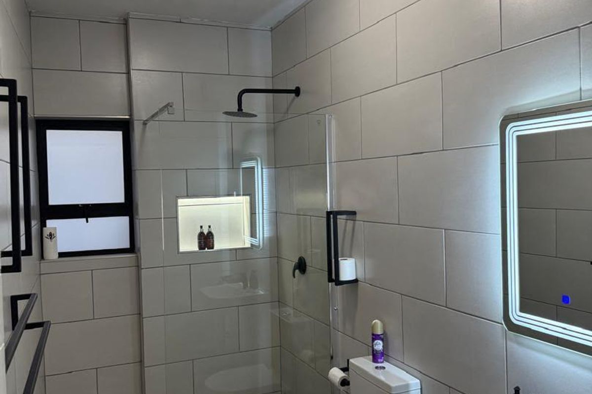 Modern bathroom featuring a walk-in shower with a recessed niche, black fixtures, an illuminated vanity mirror, and contemporary neutral tiling throughout.