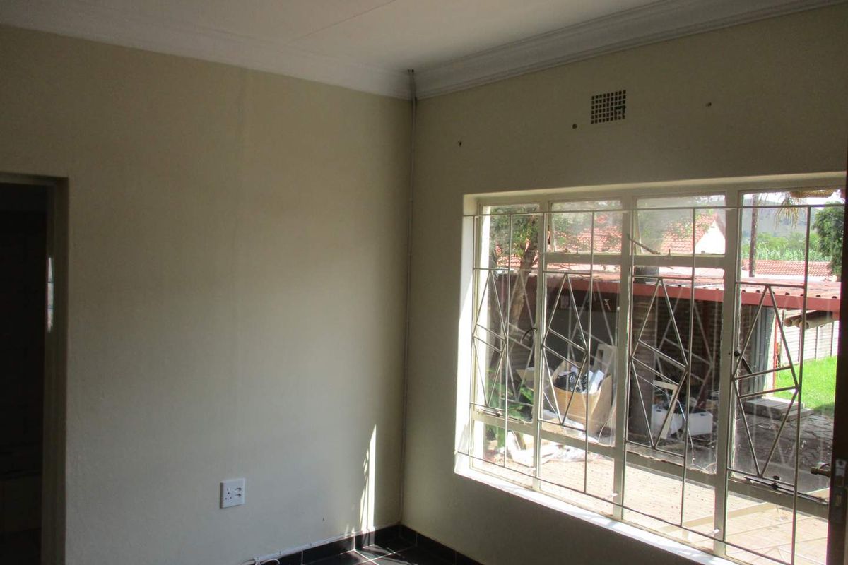 A simple, vacant room featuring dark floor tiles, neutral walls, and a large window fitted with security bars overlooking the exterior area.