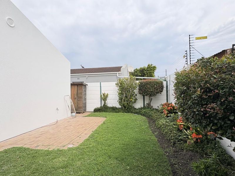 A well-maintained rear garden featuring a paved patio area, neat lawn, established garden beds, and secure boundary walling with electric fencing.