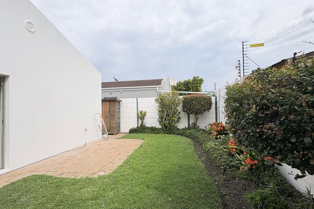 A well-maintained rear garden featuring a paved patio area, neat lawn, established garden beds, and secure boundary walling with electric fencing.