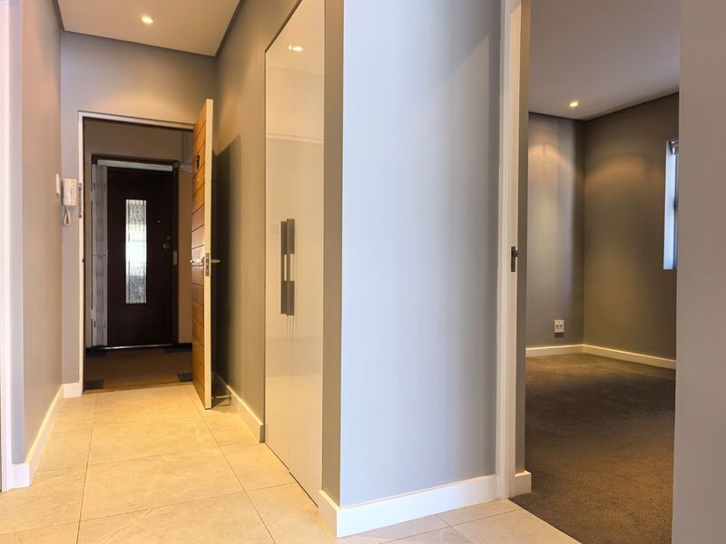 A clean, modern hallway featuring tiled flooring, a built-in cupboard with high-gloss finish, recessed lighting, and an intercom system, leading into an adjacent room.