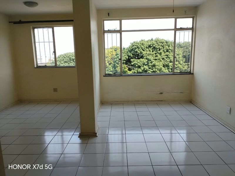 An empty living area featuring tiled flooring, large windows with security bars, and a central structural pillar, requiring minor cosmetic wall repairs.