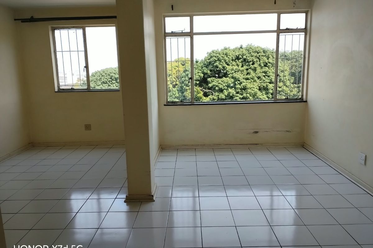 An empty living area featuring tiled flooring, large windows with security bars, and a central structural pillar, requiring minor cosmetic wall repairs.