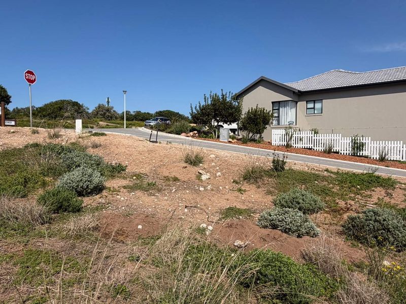 A view of the vacant residential plot showing the sloping terrain, adjacent paved driveway, and white picket fencing bordering the neighboring property.
