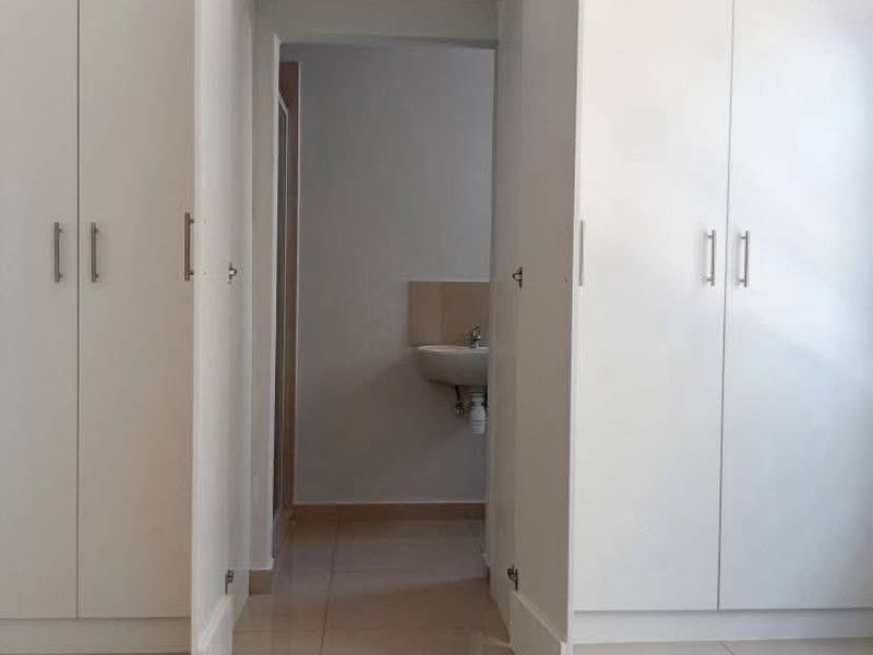 A bright bedroom featuring extensive built-in cupboards, neutral tiled flooring, and direct access to an en-suite bathroom with a wall-mounted basin.