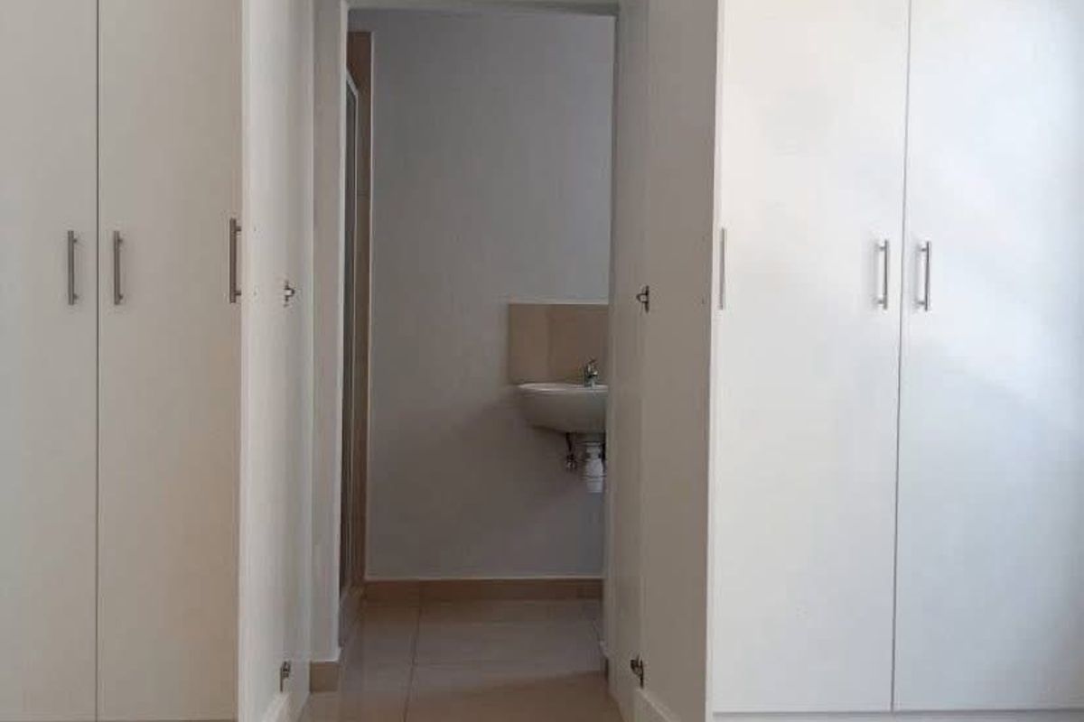 A bright bedroom featuring extensive built-in cupboards, neutral tiled flooring, and direct access to an en-suite bathroom with a wall-mounted basin.