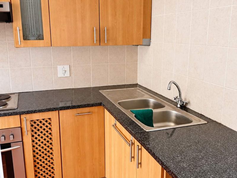 A functional kitchen featuring light wood cabinetry, dark granite countertops, a stainless steel double sink, and terracotta floor tiles.