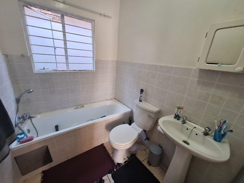 A functional bathroom featuring a bathtub, pedestal sink, and toilet, with tiled walls and a window fitted with security bars.