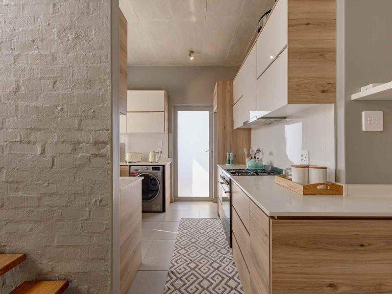 Modern kitchen featuring sleek wood-grain cabinetry, stone countertops, gas hob, and an exposed brick wall, leading to a rear exterior door.