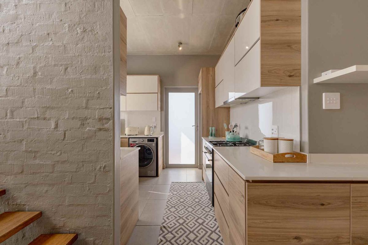 Modern kitchen featuring sleek wood-grain cabinetry, stone countertops, gas hob, and an exposed brick wall, leading to a rear exterior door.