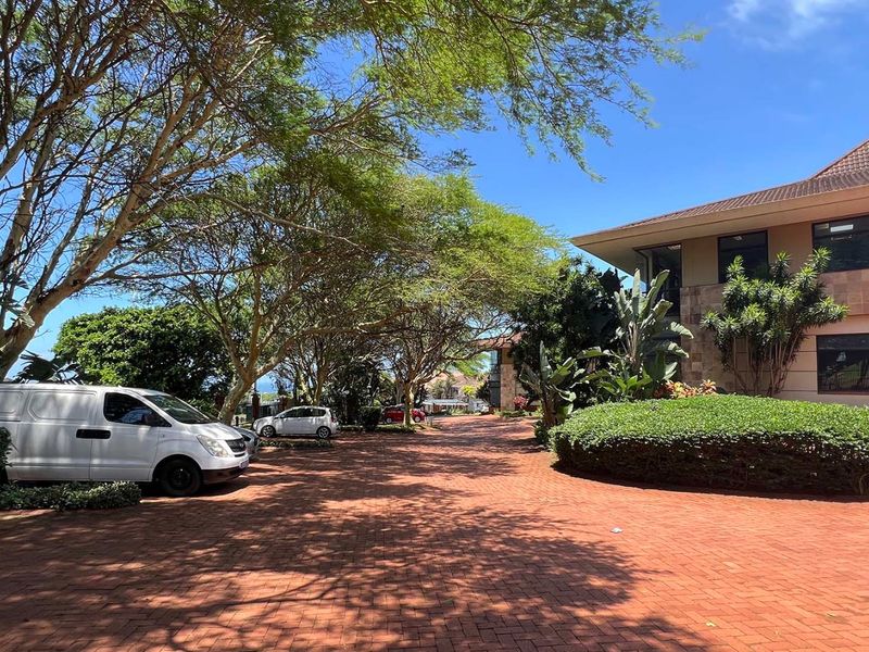 A well-maintained office property featuring a paved driveway, mature trees, landscaped garden areas, and a multi-story building facade with a tiled roof.