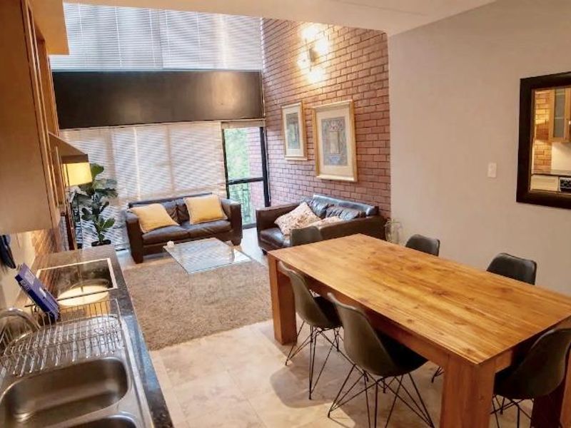 Open-plan living and dining area featuring an exposed brick wall, tiled flooring, and a functional kitchen with double sink and cabinetry.