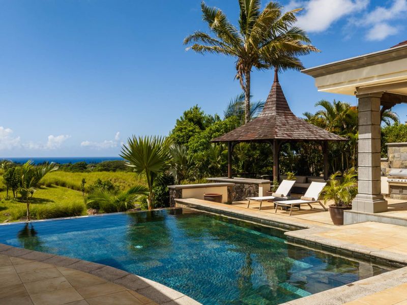 A stunning outdoor pool area featuring a rim-flow swimming pool, stone-tiled patio, and a covered gazebo, set against a scenic landscape and ocean view.