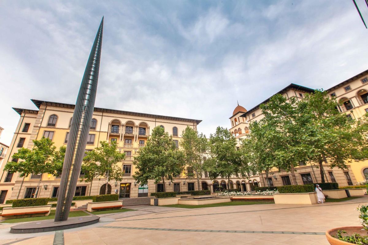 A well-maintained office complex courtyard featuring paved walkways, mature trees, landscaped garden beds, and a prominent sculptural spire.