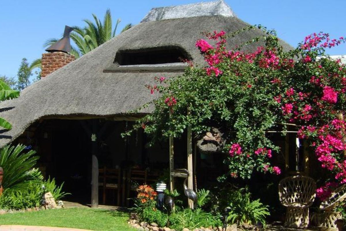 A charming thatched-roof guest house structure featuring a brick chimney and a covered patio area, surrounded by lush garden landscaping.