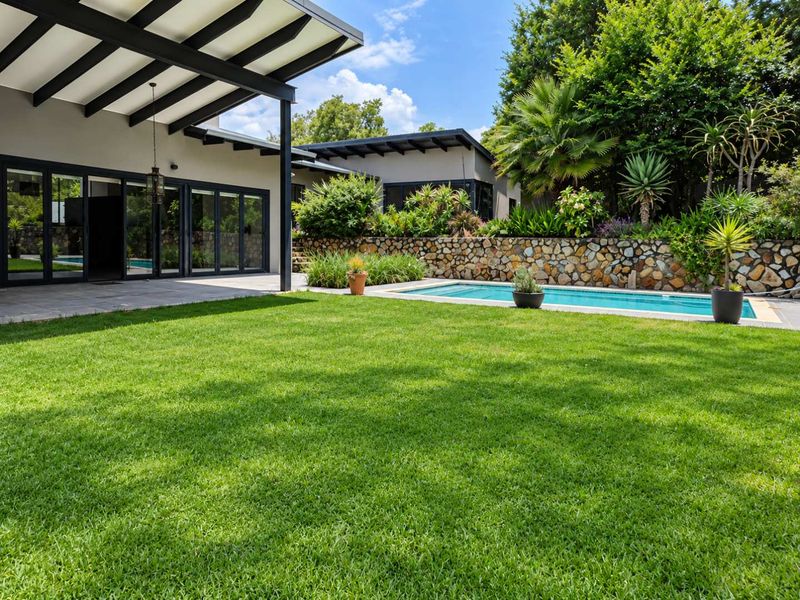 A well-maintained backyard featuring a swimming pool, stone retaining wall, lush lawn, and a covered patio area with modern bi-fold doors.
