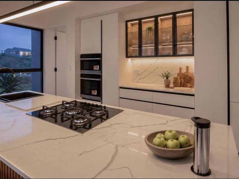 Modern kitchen featuring a large stone island with gas cooktop, integrated double ovens, and elegant glass-fronted display cabinetry with ambient lighting.