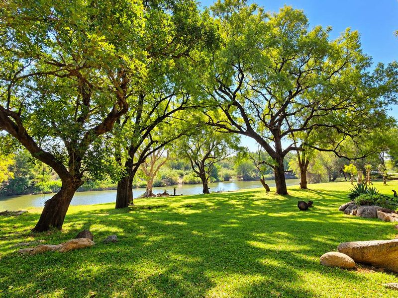 A beautifully maintained riverside garden featuring mature trees, expansive green lawns, and natural rock features, offering a serene outdoor setting.