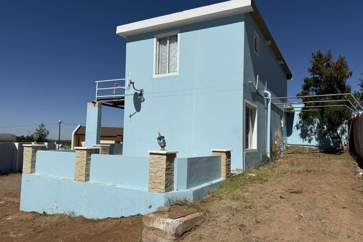 Rear view of a two-story house featuring a low boundary wall with stone-clad pillars, a small balcony, and an unlandscaped yard area.
