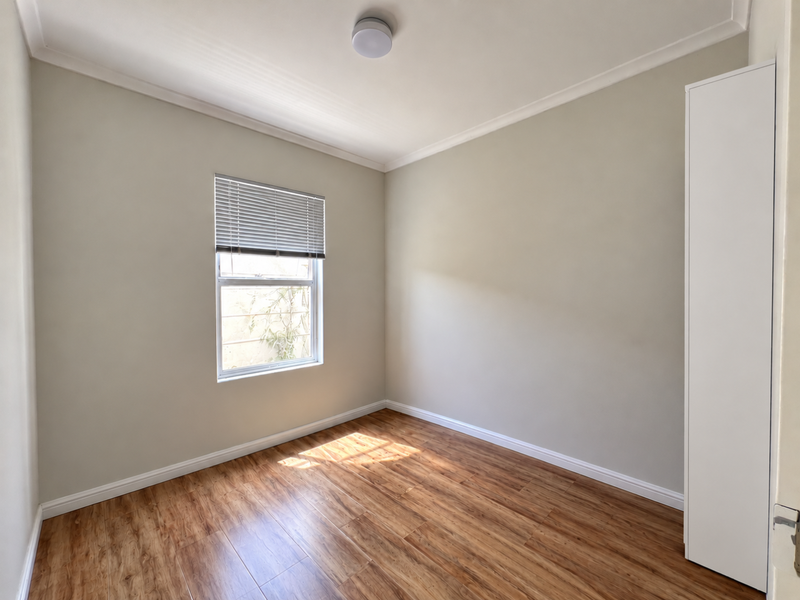 A bright and neutral bedroom featuring laminate flooring, a built-in cupboard, and a window with a fitted venetian blind.