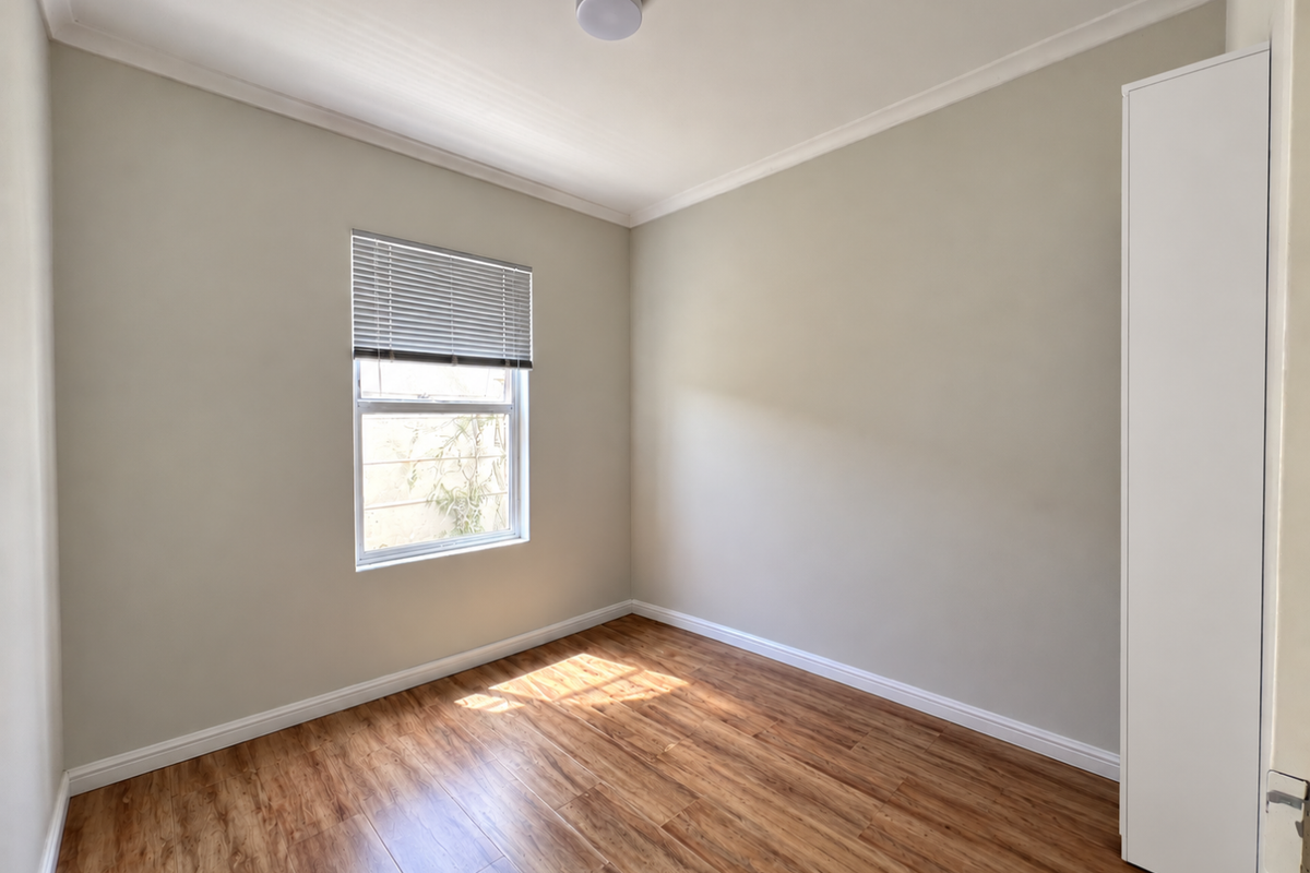 A bright and neutral bedroom featuring laminate flooring, a built-in cupboard, and a window with a fitted venetian blind.