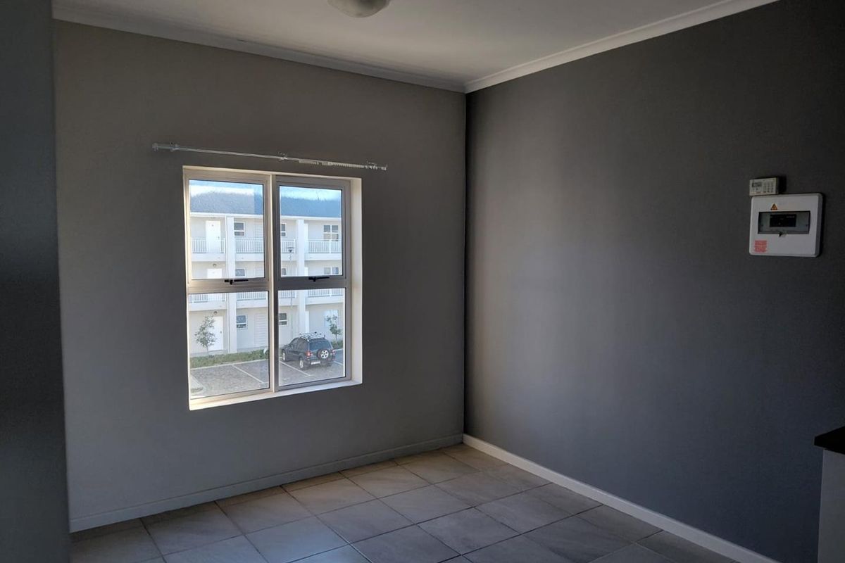 An empty, neutrally painted living area featuring tiled flooring, a ceiling light, and a window overlooking the apartment complex parking area.