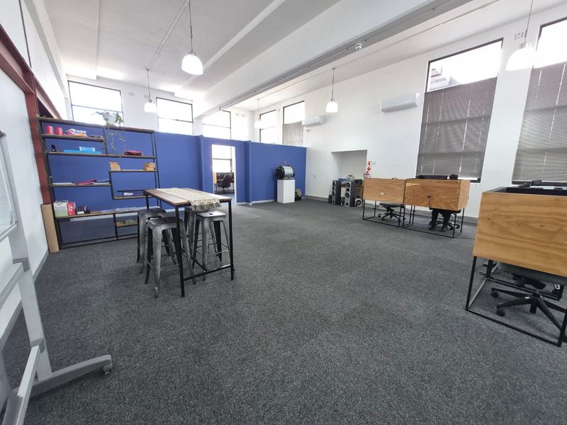 Spacious open-plan office area featuring high ceilings, exposed structural elements, large windows for natural light, and modern carpeted flooring.