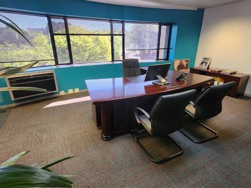 A functional office space featuring large windows with venetian blinds, wall-to-wall carpeting, and a built-in air conditioning unit.