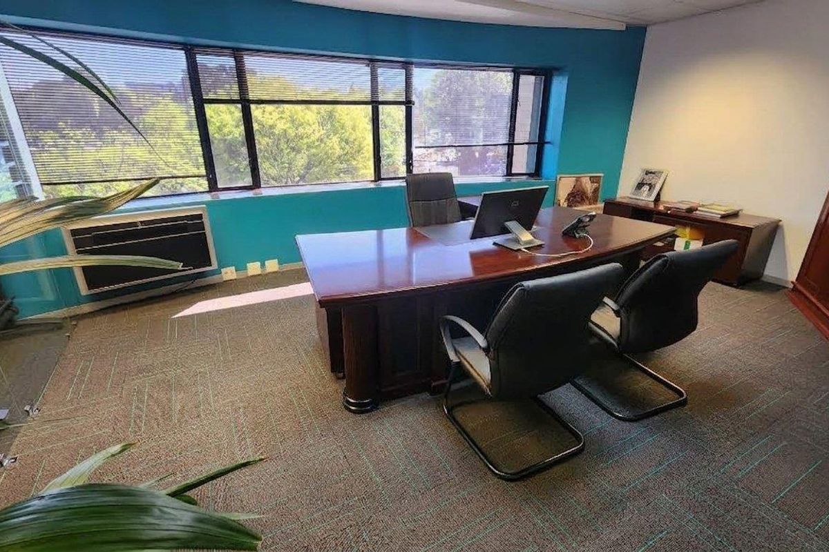 A functional office space featuring large windows with venetian blinds, wall-to-wall carpeting, and a built-in air conditioning unit.