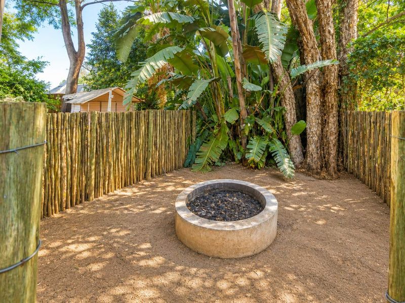 A private outdoor garden area featuring a circular concrete boma fire pit enclosed by rustic wooden pole fencing and surrounded by mature trees.