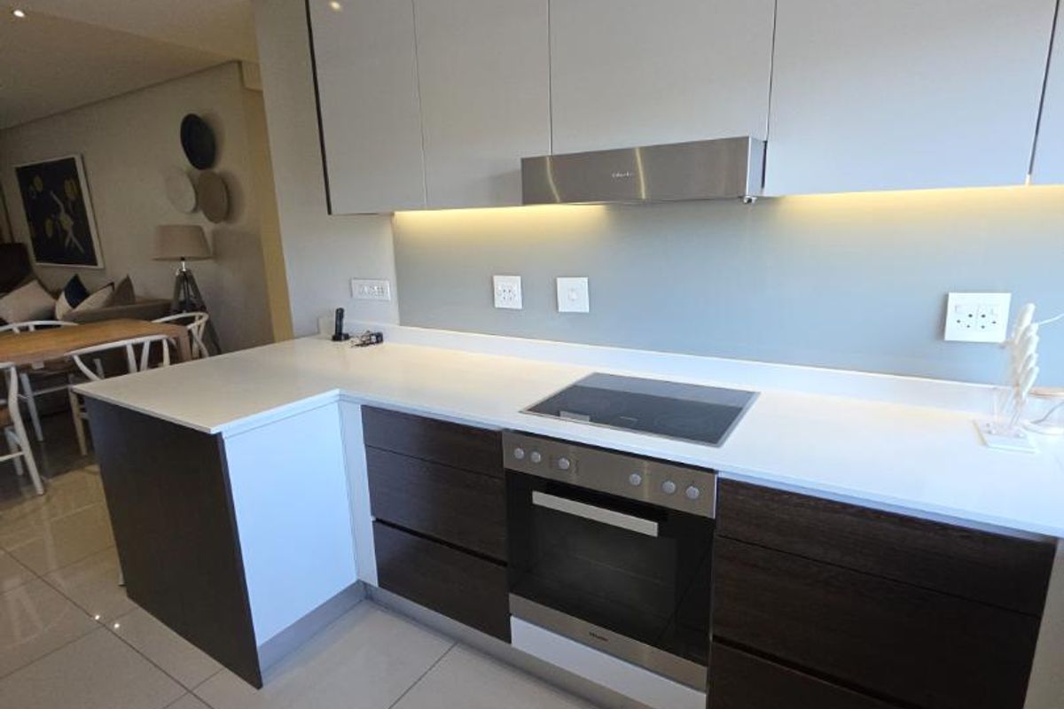 Modern kitchen featuring sleek two-tone cabinetry, stone countertops, integrated oven and hob, and ambient under-cabinet lighting, seamlessly flowing into the adjacent living area.