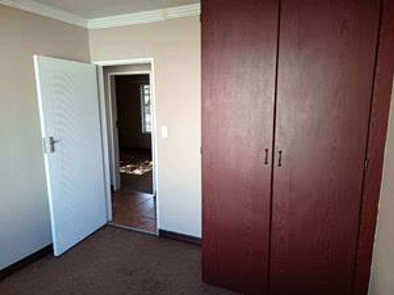 A vacant bedroom featuring built-in wooden wardrobes, carpeted flooring, and neutral wall finishes, with an adjacent hallway visible through the doorway.