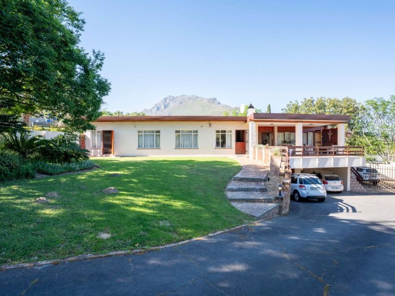 Front exterior view of a single-story residence featuring a paved driveway, stone staircase, raised patio area, and undercover parking with mountain views.