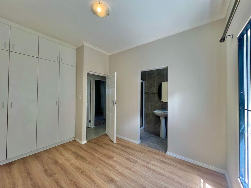 A bright, empty bedroom featuring built-in cupboards, laminate flooring, and direct access to an en-suite bathroom with a pedestal basin.