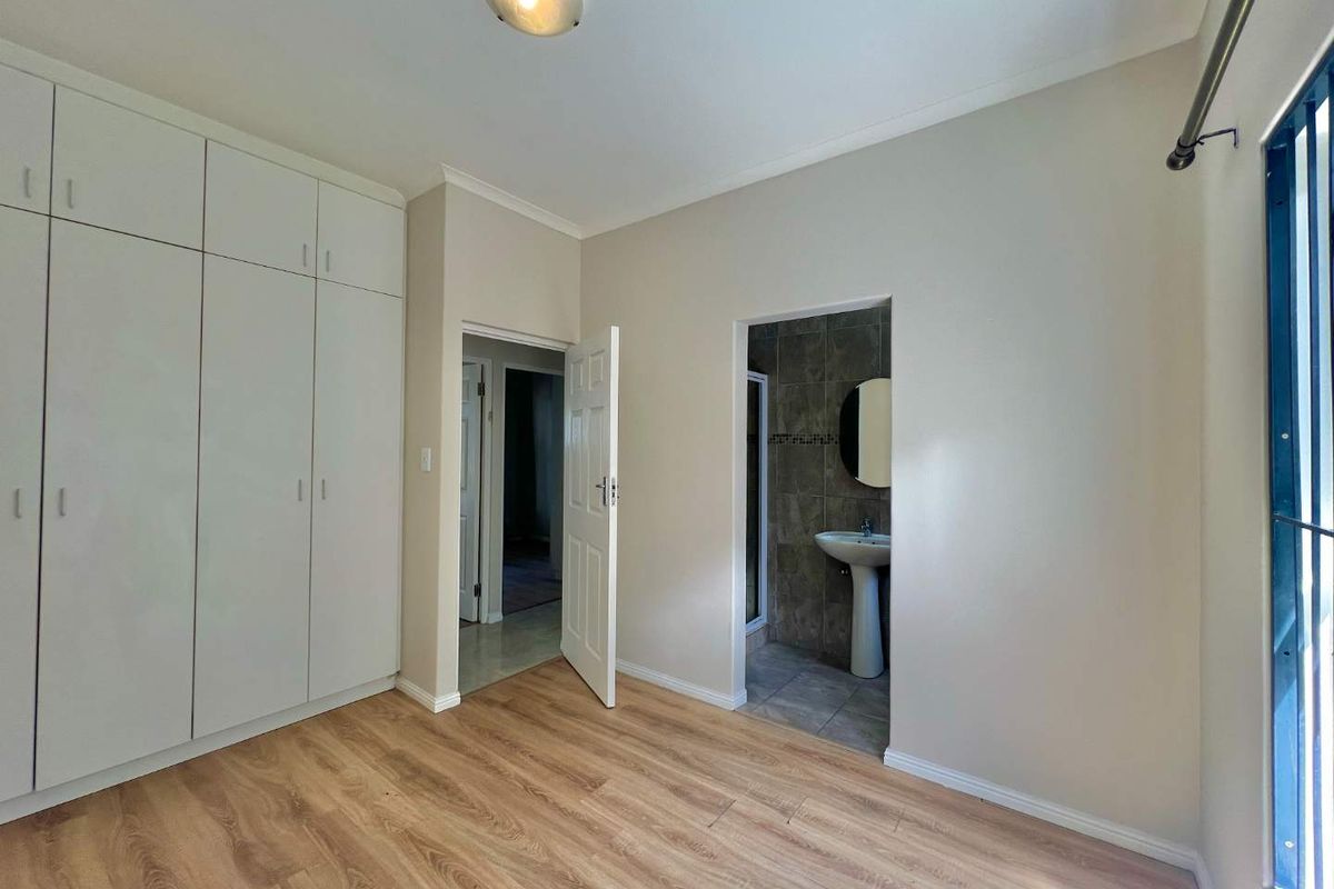 A bright, empty bedroom featuring built-in cupboards, laminate flooring, and direct access to an en-suite bathroom with a pedestal basin.