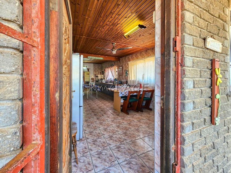 A dated kitchen featuring timber ceilings, ceramic tiled flooring, and built-in cabinetry, viewed from an exterior doorway with exposed brick walls.