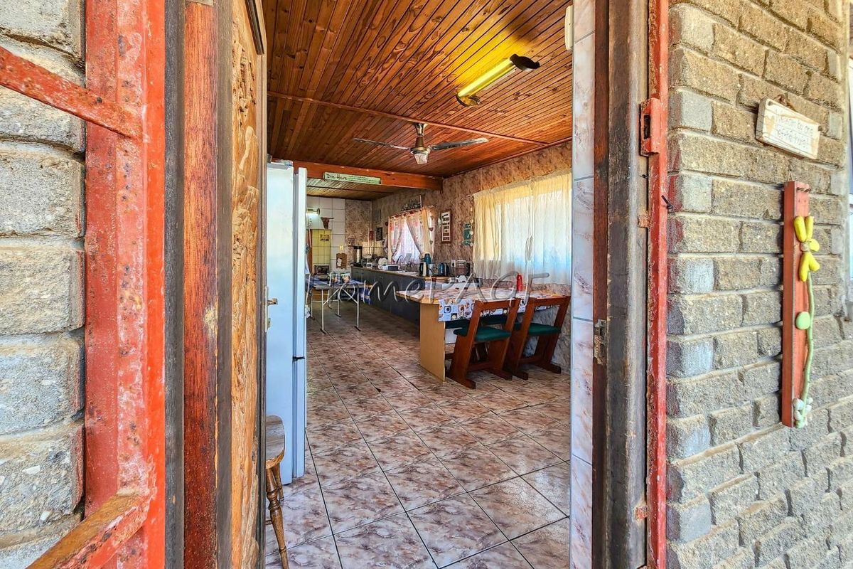 A dated kitchen featuring timber ceilings, ceramic tiled flooring, and built-in cabinetry, viewed from an exterior doorway with exposed brick walls.