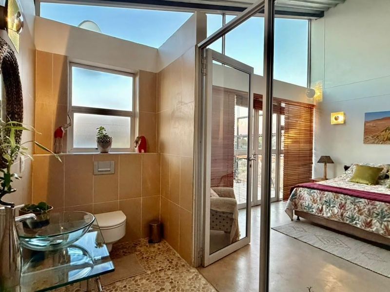Modern bedroom featuring an open-plan en-suite bathroom with a glass vessel sink, wall-mounted toilet, and floor-to-ceiling windows providing ample natural light.