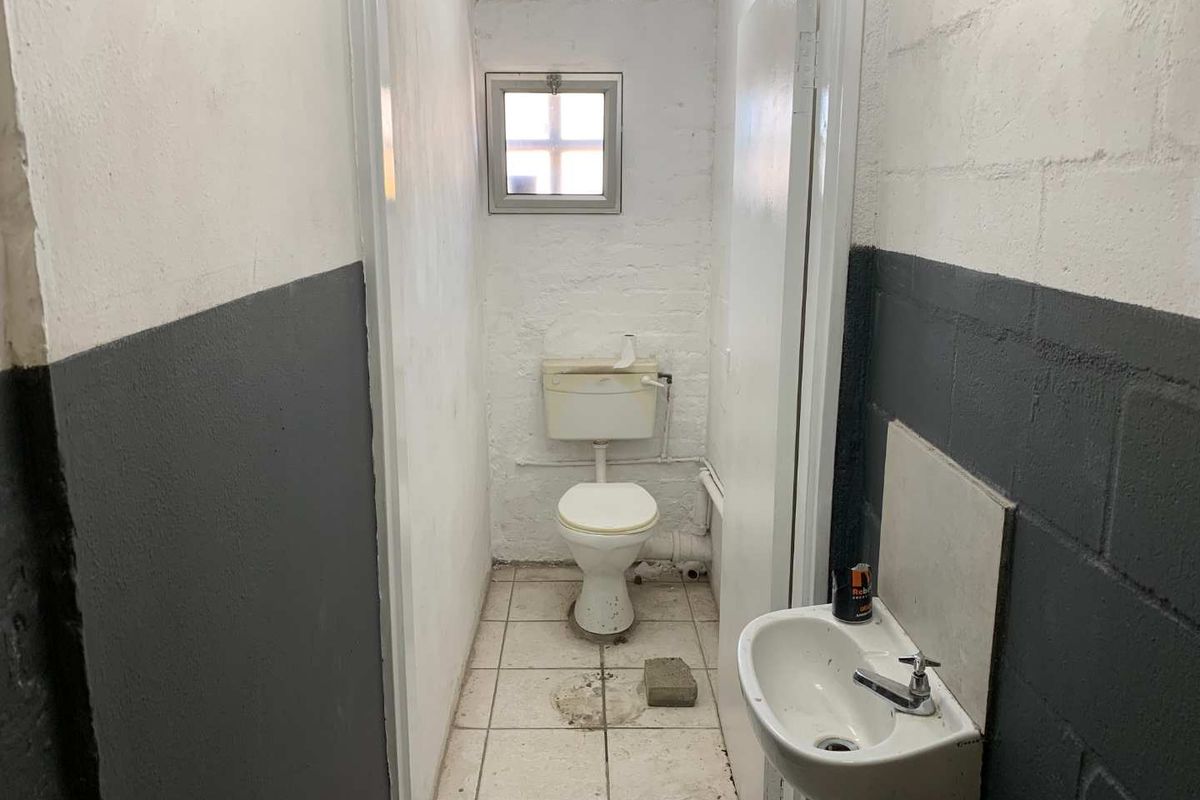 A small, utilitarian industrial restroom featuring a basic toilet, wall-mounted basin, and tiled flooring with exposed brick walls.