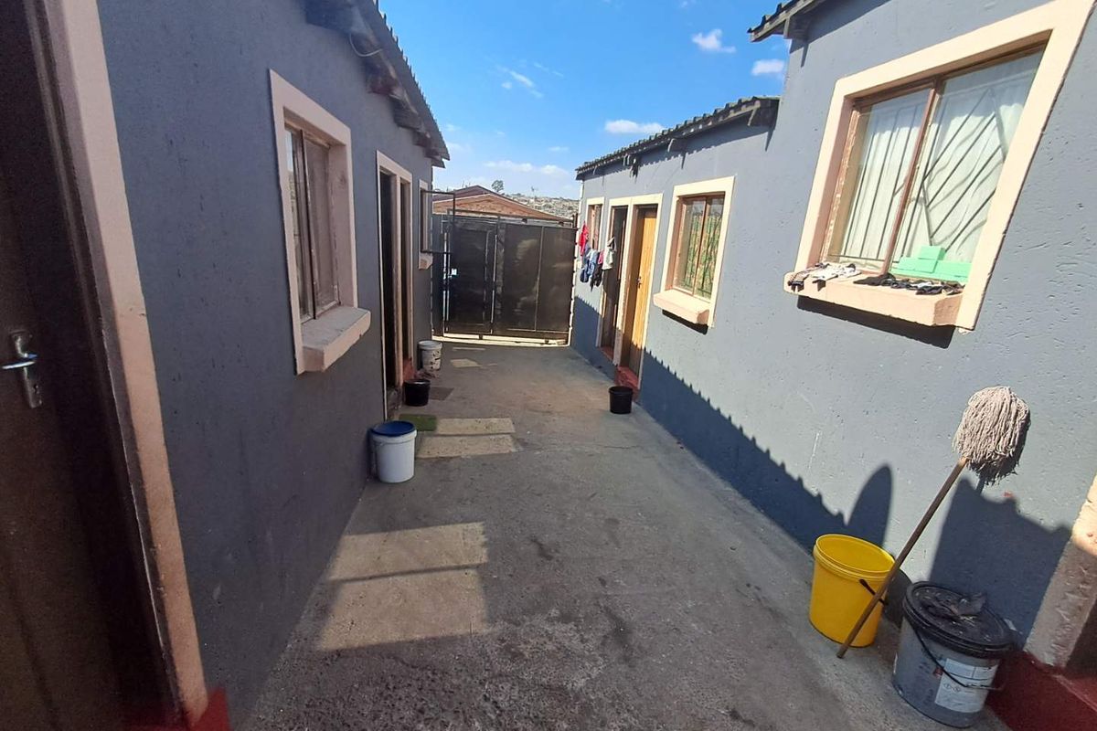 A narrow exterior walkway between two residential structures featuring concrete paving, security bars on windows, and a metal access gate.