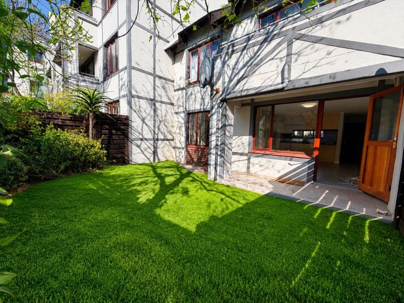 A private rear garden featuring a well-maintained lawn, timber-framed windows, and a paved patio area providing direct access to the apartment interior.