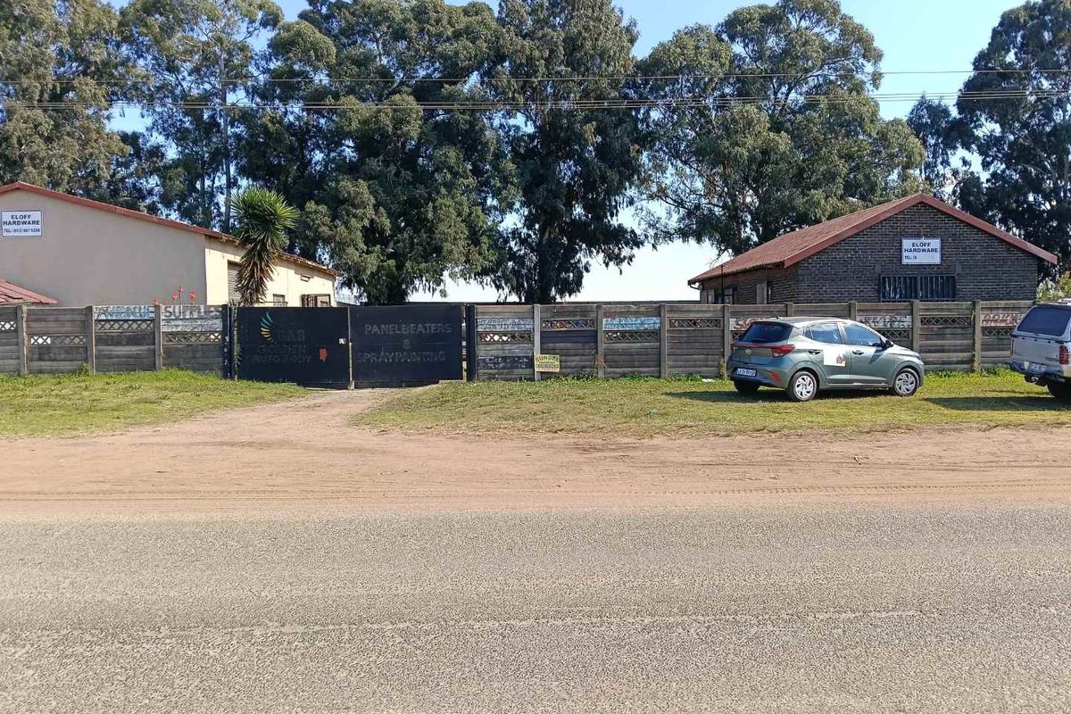 Street view of a commercial property featuring two brick buildings, a gated entrance, and perimeter fencing, situated along an unpaved road.