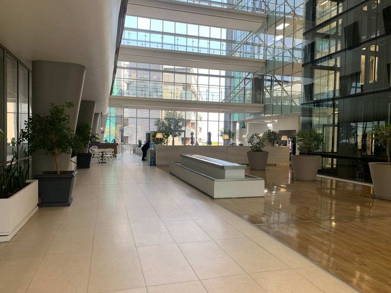 Spacious modern office building atrium featuring high ceilings, extensive glass curtain walls, and a combination of tiled and wood-look flooring.