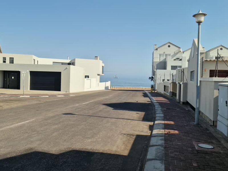 A clear street view showing the neighbourhood approach with paved sidewalks, boundary walls, and a distant view of the ocean.