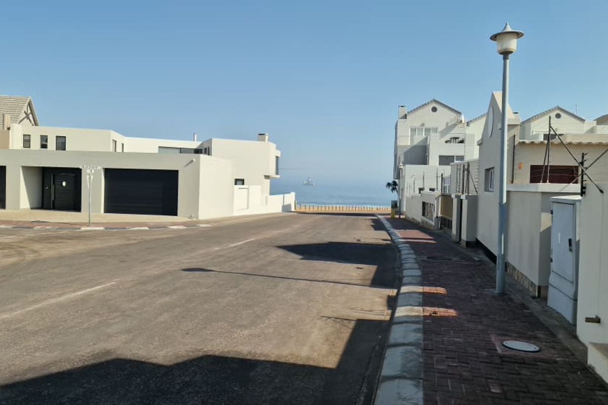 A clear street view showing the neighbourhood approach with paved sidewalks, boundary walls, and a distant view of the ocean.