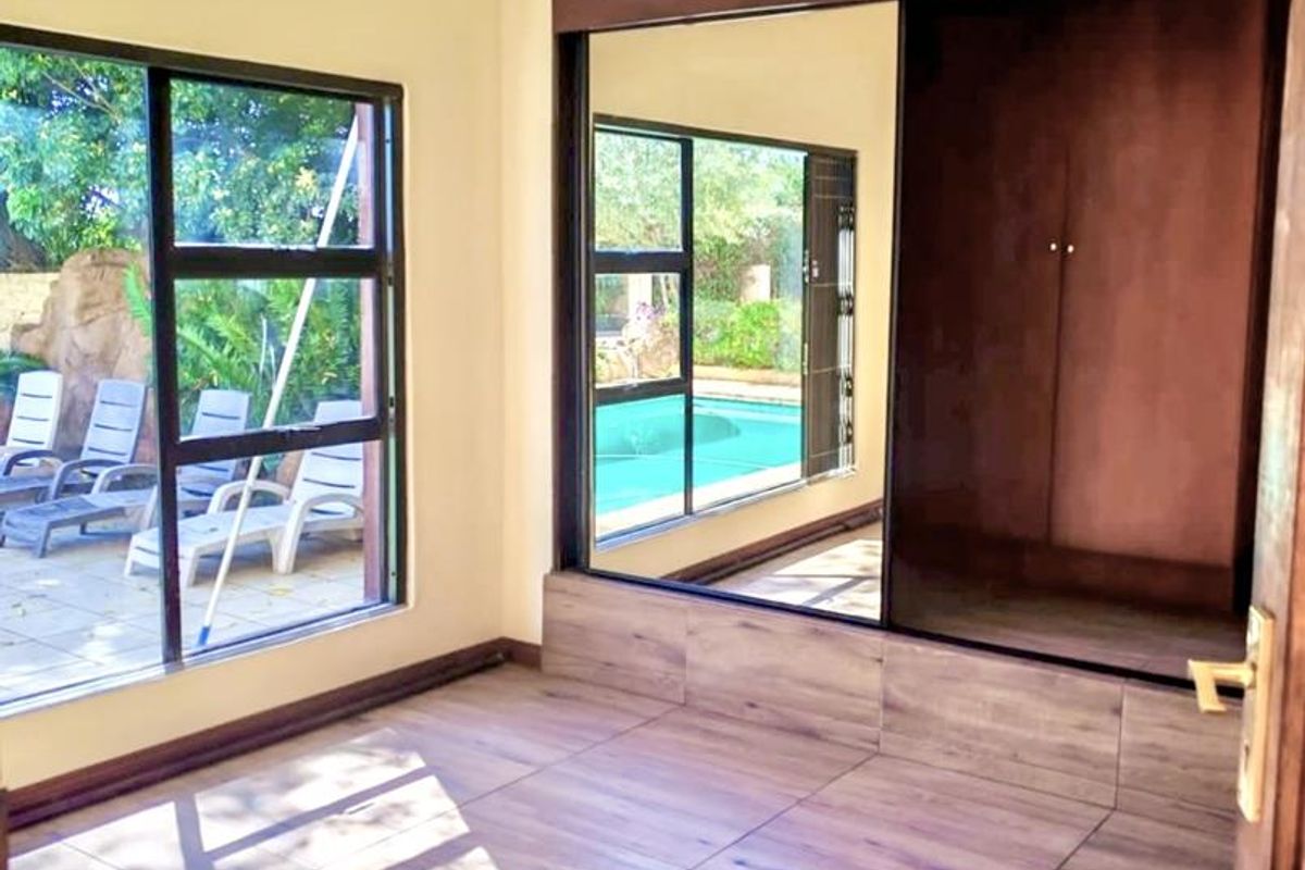 A bright, empty bedroom featuring wood-look tiled flooring, a ceiling fan, and built-in cupboards with mirrored doors, offering views of the pool area.