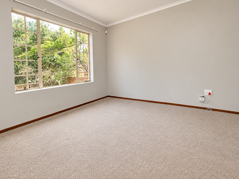 A bright, empty bedroom featuring neutral walls, carpeted flooring, timber skirting boards, and a large window overlooking the garden.