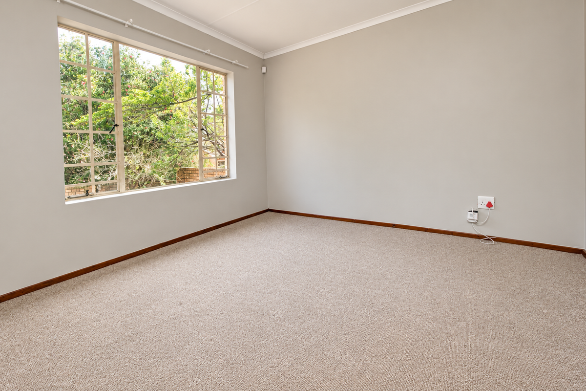 A bright, empty bedroom featuring neutral walls, carpeted flooring, timber skirting boards, and a large window overlooking the garden.
