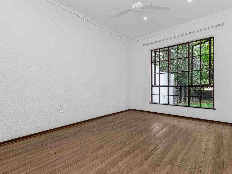 A bright, empty bedroom featuring modern laminate flooring, a ceiling fan, and a large window overlooking the garden.